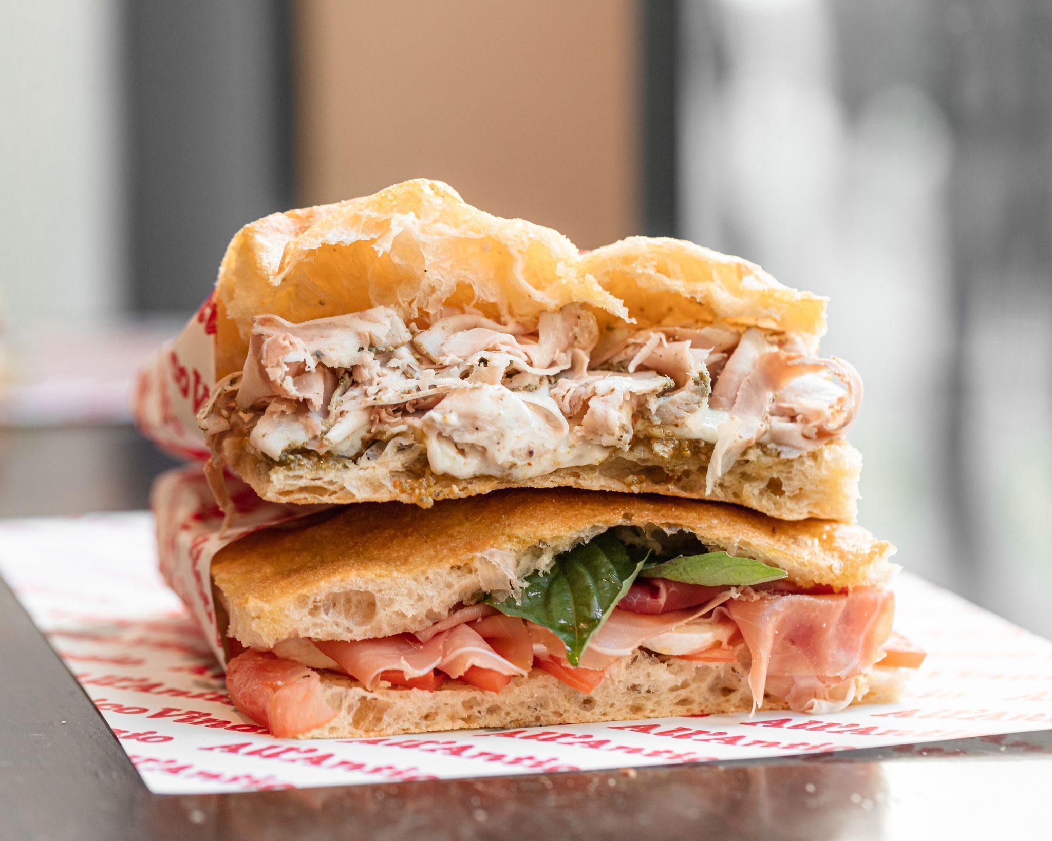A close-up of a thick, Italian sandwich from All'Antico Vinaio with a crisp flatbread base, sliced prosciutto, fresh basil, and what appears to be a second layer of bread with shredded meat and a white sauce.