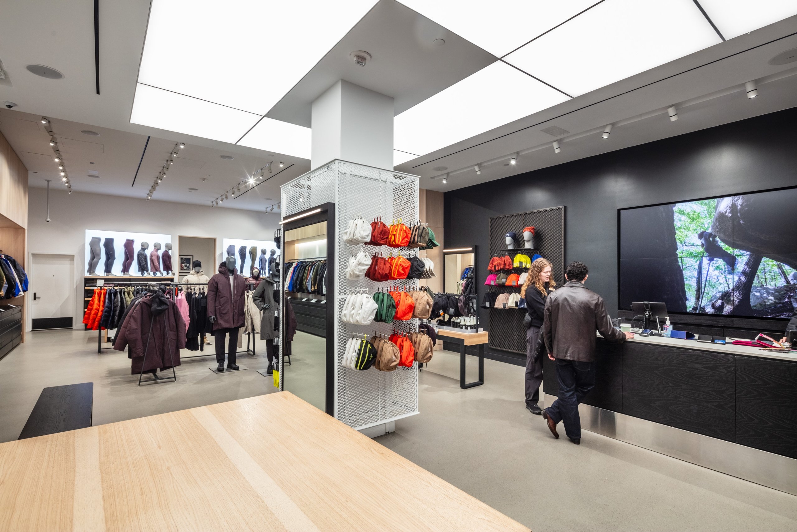 The interior of an Arc'teryx outdoor clothing store with a modern design, featuring black walls, large video screens, a checkout counter, and a white mesh display rack showcasing colorful small backpacks and gear.
