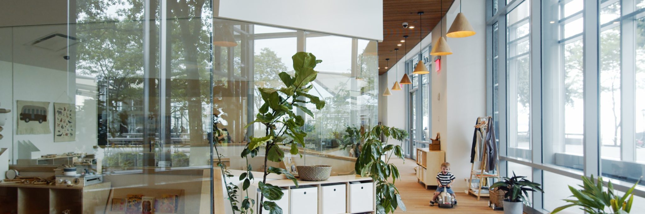 The brightly lit interior of a Big and Tiny children's play space with floor-to-ceiling windows overlooking an outdoor area. A young child is riding a toy vehicle on a light wood floor surrounded by potted plants and a toy shelf.