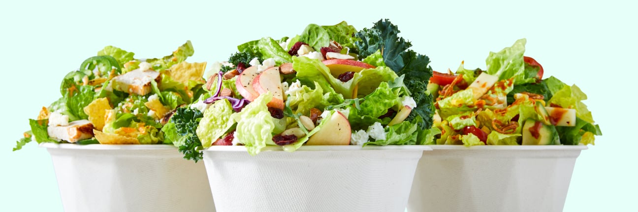 A close-up, stylized view of three large salads in compostable white bowls from Chopt, showing mounds of fresh green lettuce, kale, and various toppings.