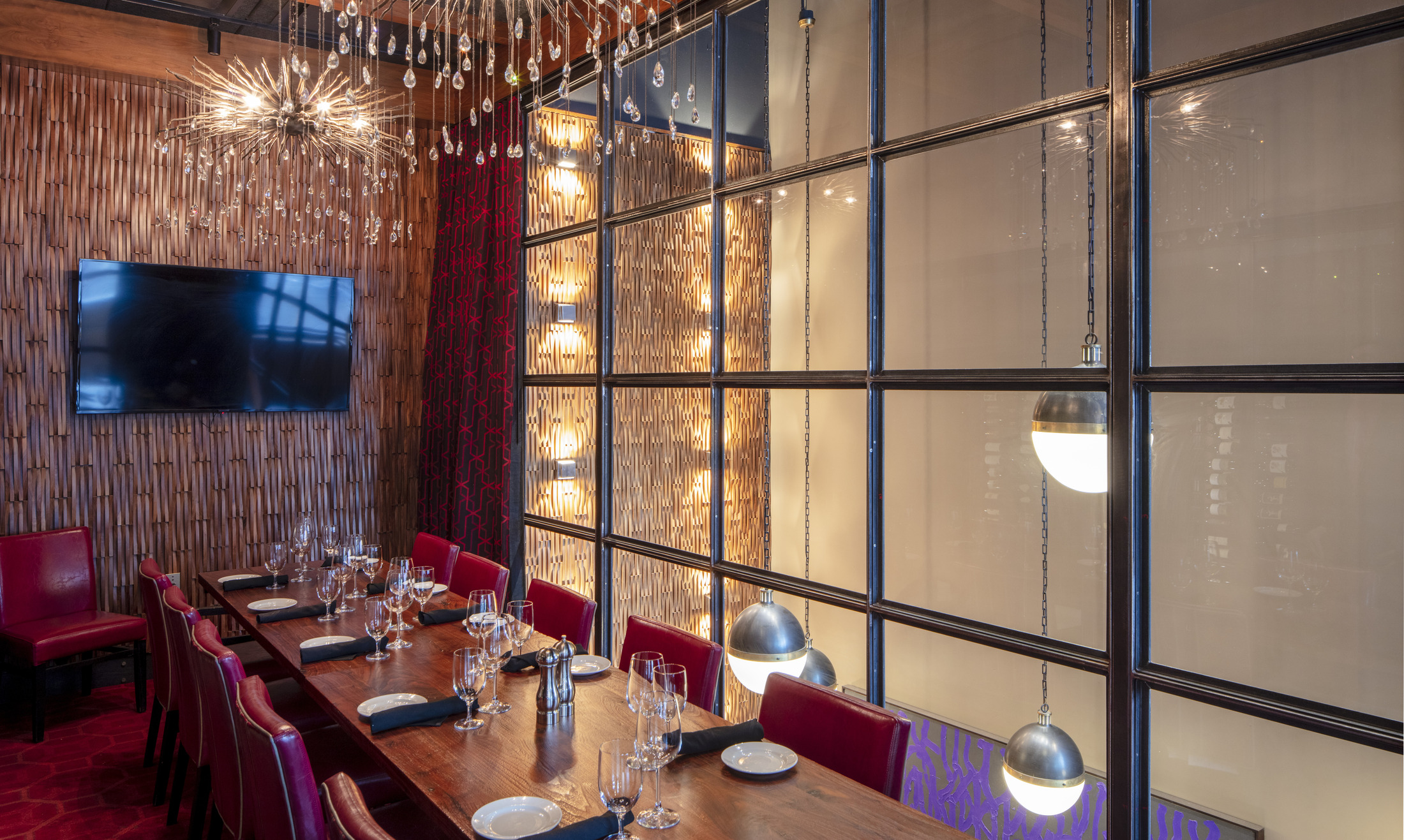 A view of a private dining room in a restaurant with a long wooden table, red leather chairs, a flat-screen TV, a textured wooden wall, and large, ornate crystal chandeliers.