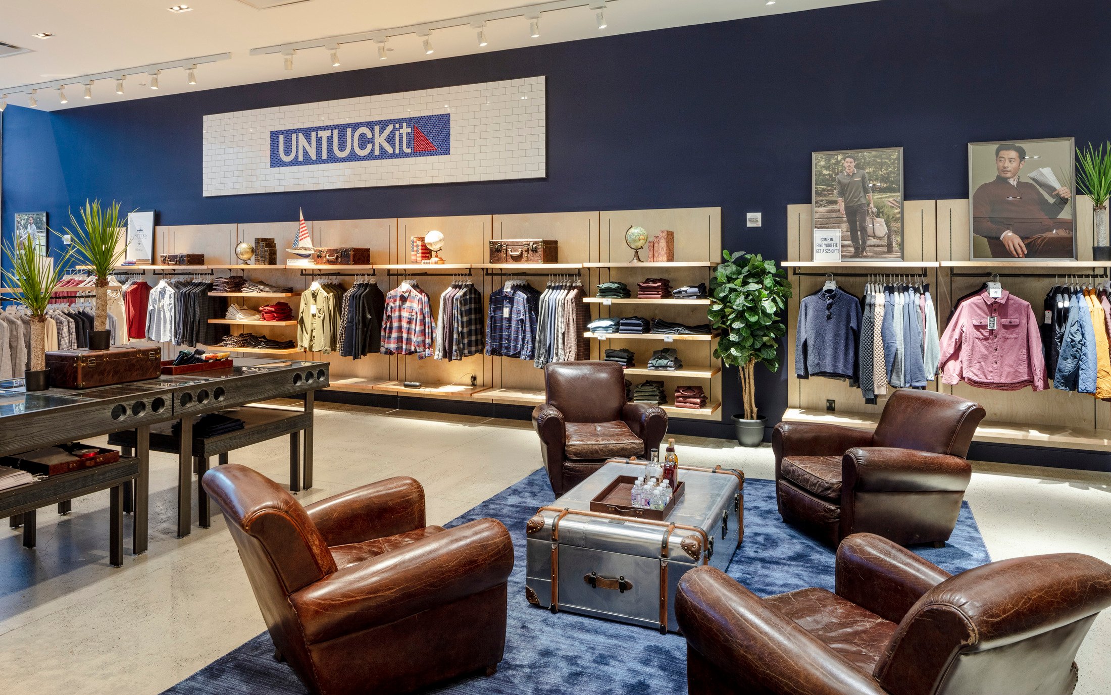 Interior of a men's clothing store with casual furniture, wooden accents, and stacked apparel displays.