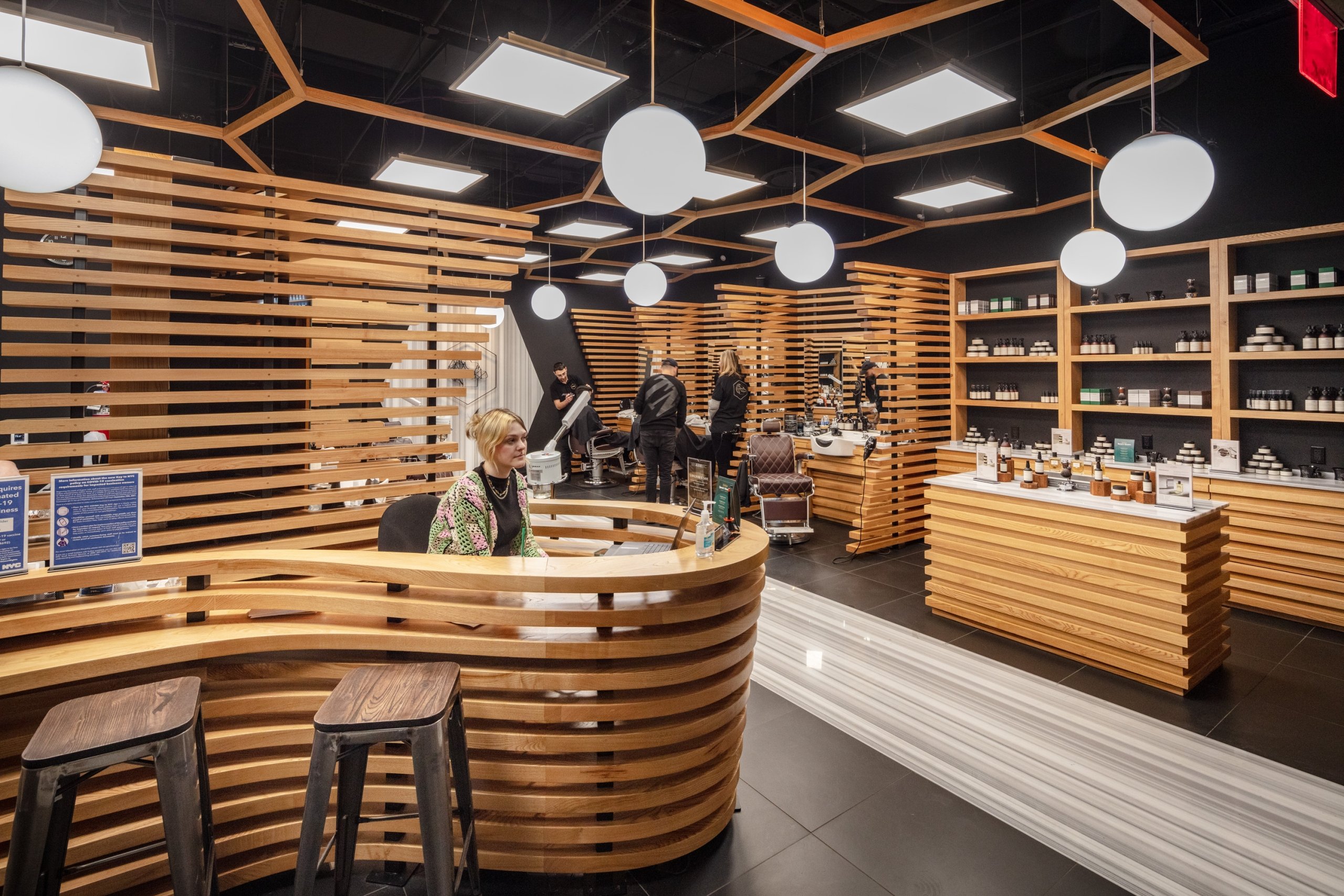 The interior of a modern grooming shop features a curvilinear, slatted wood reception desk and matching wooden walls, with a black ceiling, globe lighting, and black shelves stocked with products.