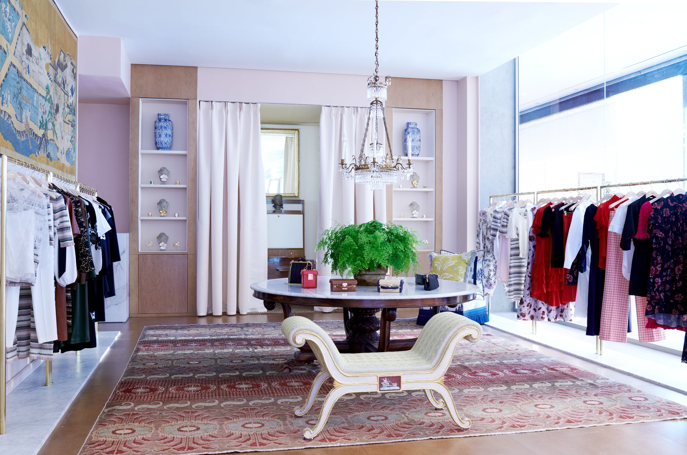 The interior of an Adam Lippes clothing boutique with pale pink walls, a red patterned rug, and gold-trimmed racks of clothing. A marble-topped table and a crystal chandelier furnish the center of the room.