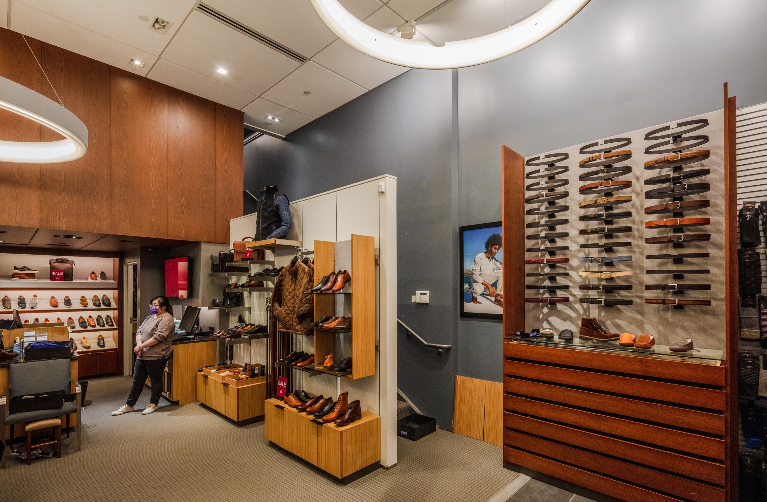 The interior of an Allen Edmonds shoe store, showcasing leather shoes and belts on tiered wooden and glass display cases against a backdrop of dark gray and wood-paneled walls.