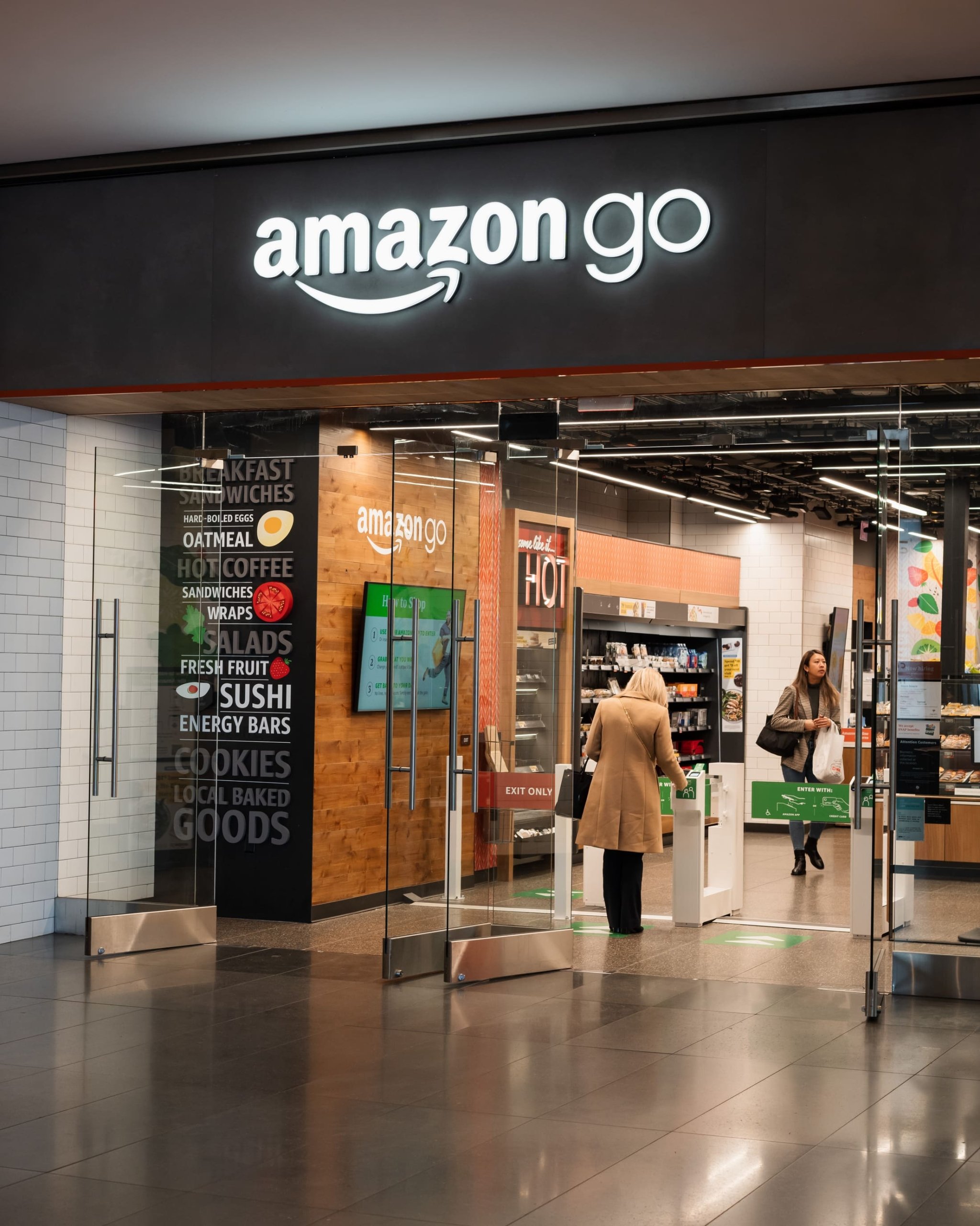 The entrance to an Amazon Go store with glass doors. The interior shows food displays and a woman exiting, with a vertical sign listing food items like