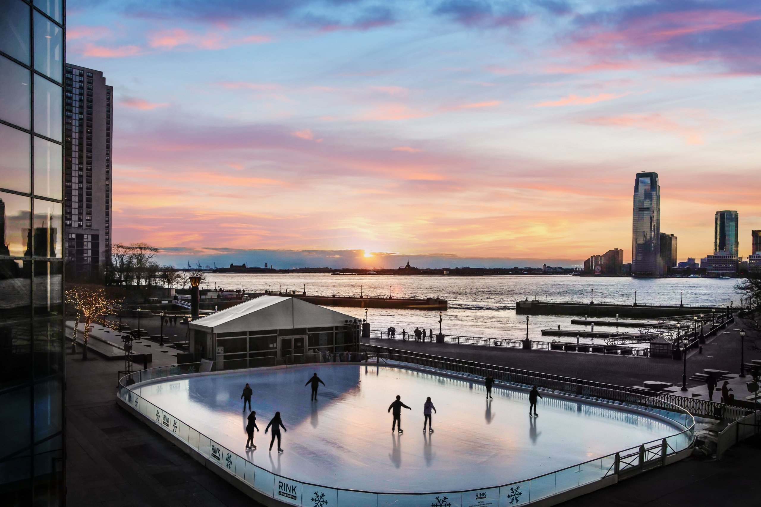 Outdoor ice rink with a few skaters at sunset, overlooking a waterfront and city skyline in the distance.