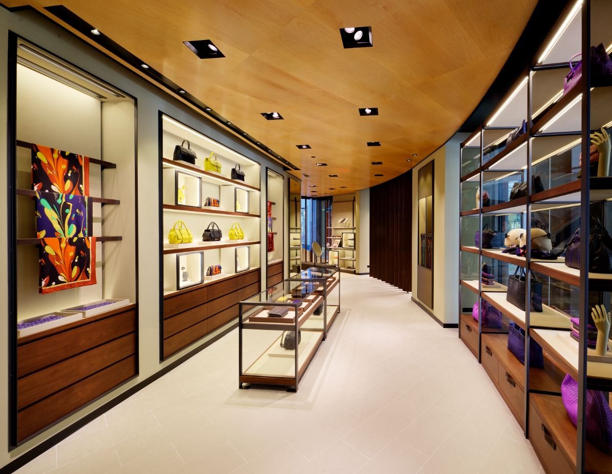 The interior of a luxury Bottega Veneta boutique with a light wood-paneled ceiling, cream-colored tile floor, and elegant display shelves showcasing handbags and accessories in bright and muted colors.