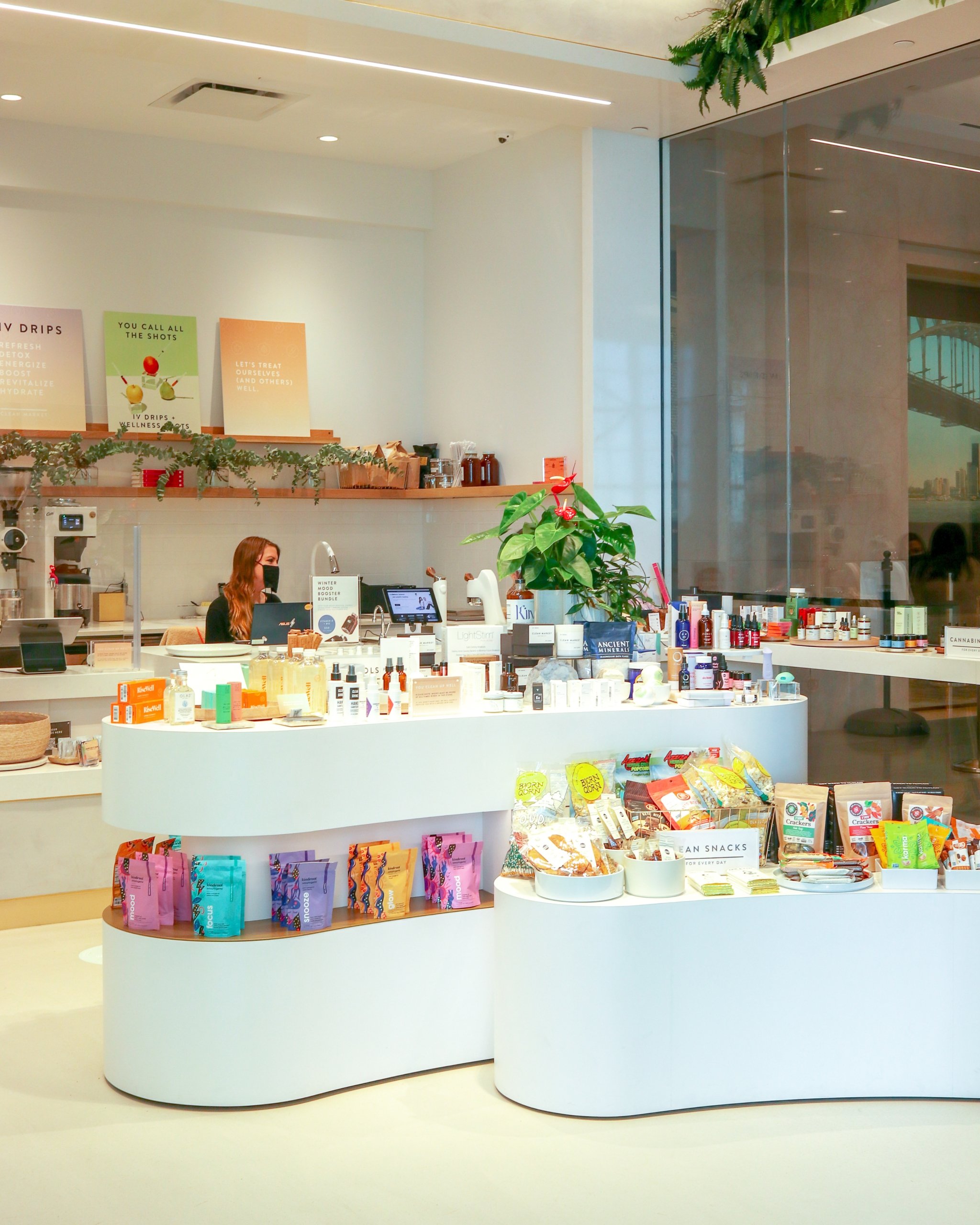 A brightly lit, modern health and wellness store with white counters displaying colorful packages of snacks and health products. A person works behind the main counter with a selection of IV drip menu posters visible above.
