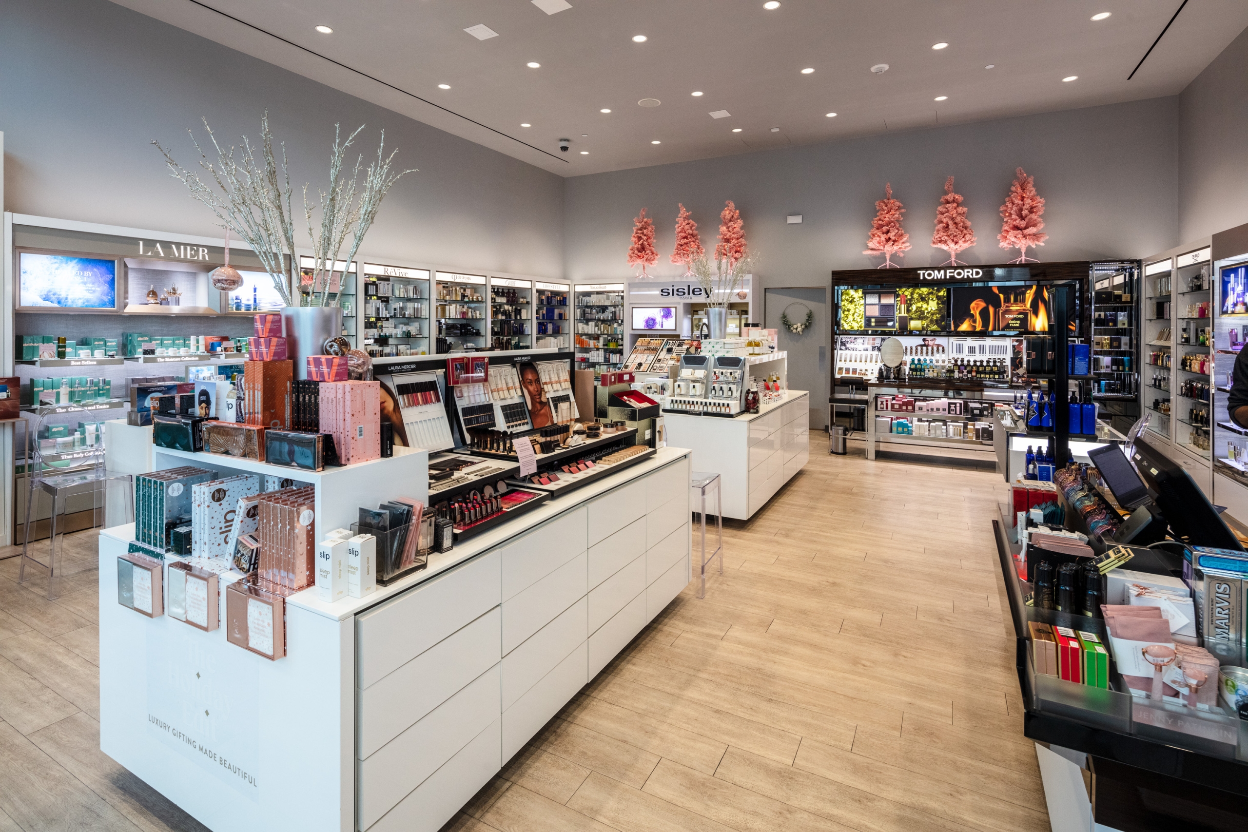 The clean, modern interior of a Cos Bar cosmetics store, featuring bright white display islands and sleek shelves stocked with luxury makeup and skincare products, with subtle pink decorations in the background.