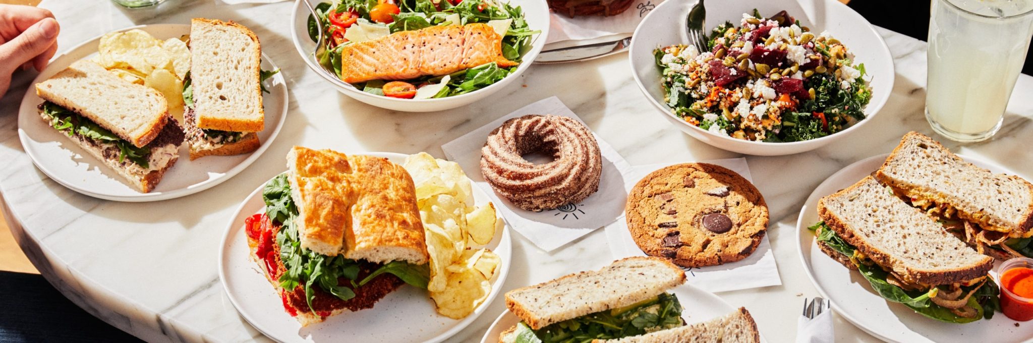 A marble tabletop covered with a variety of lunch items, including salmon salad, several sandwiches, potato chips, a cinnamon swirl pastry, and a large chocolate chip cookie.