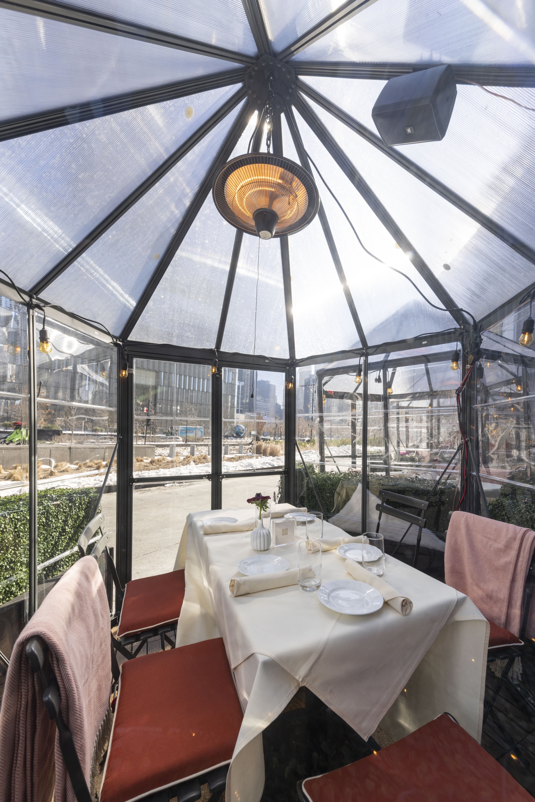 Outdoor dining area set up inside a glass-walled greenhouse or pavilion with white furniture.