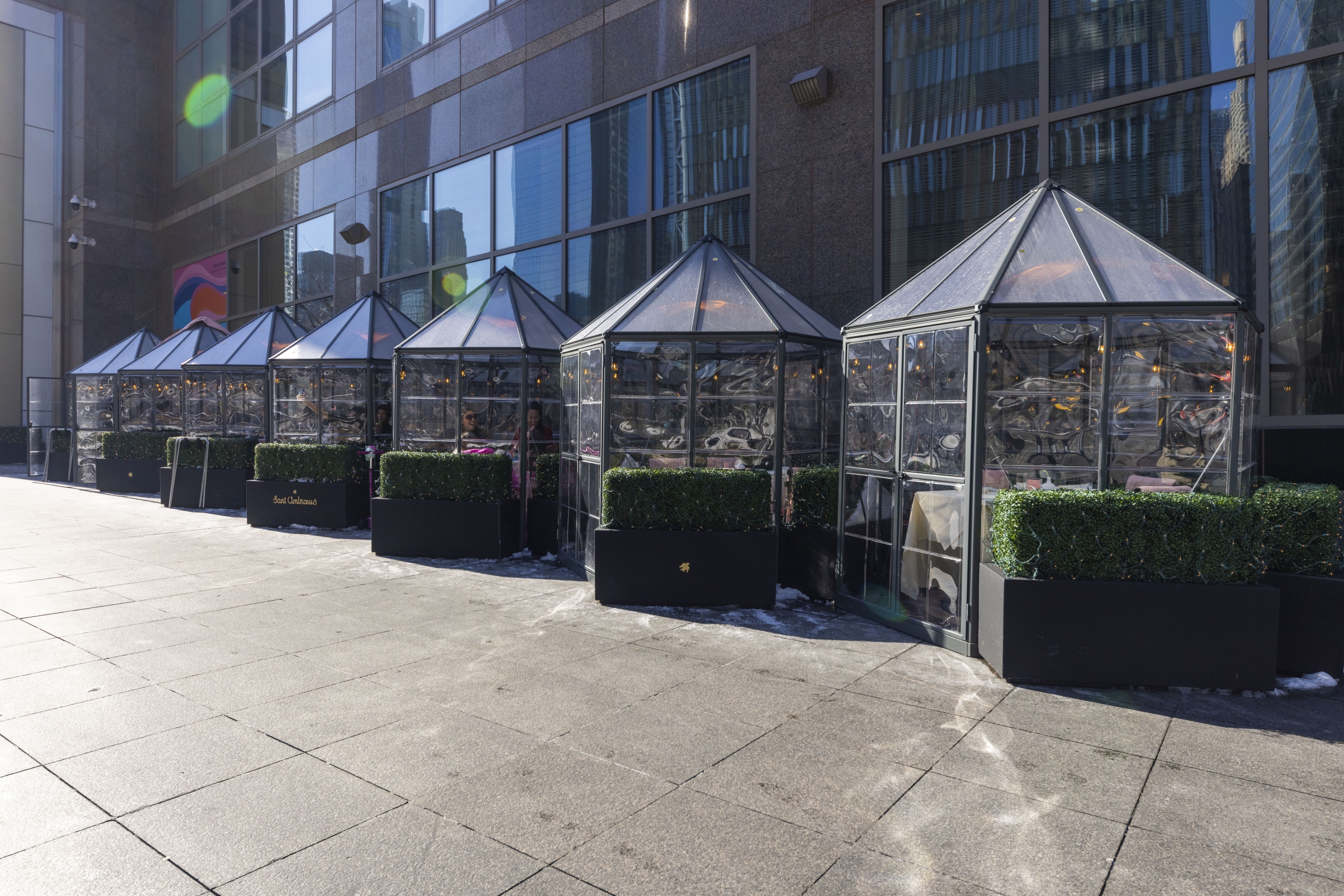 Row of semi-private outdoor dining booths with glass walls and transparent plastic roofs.