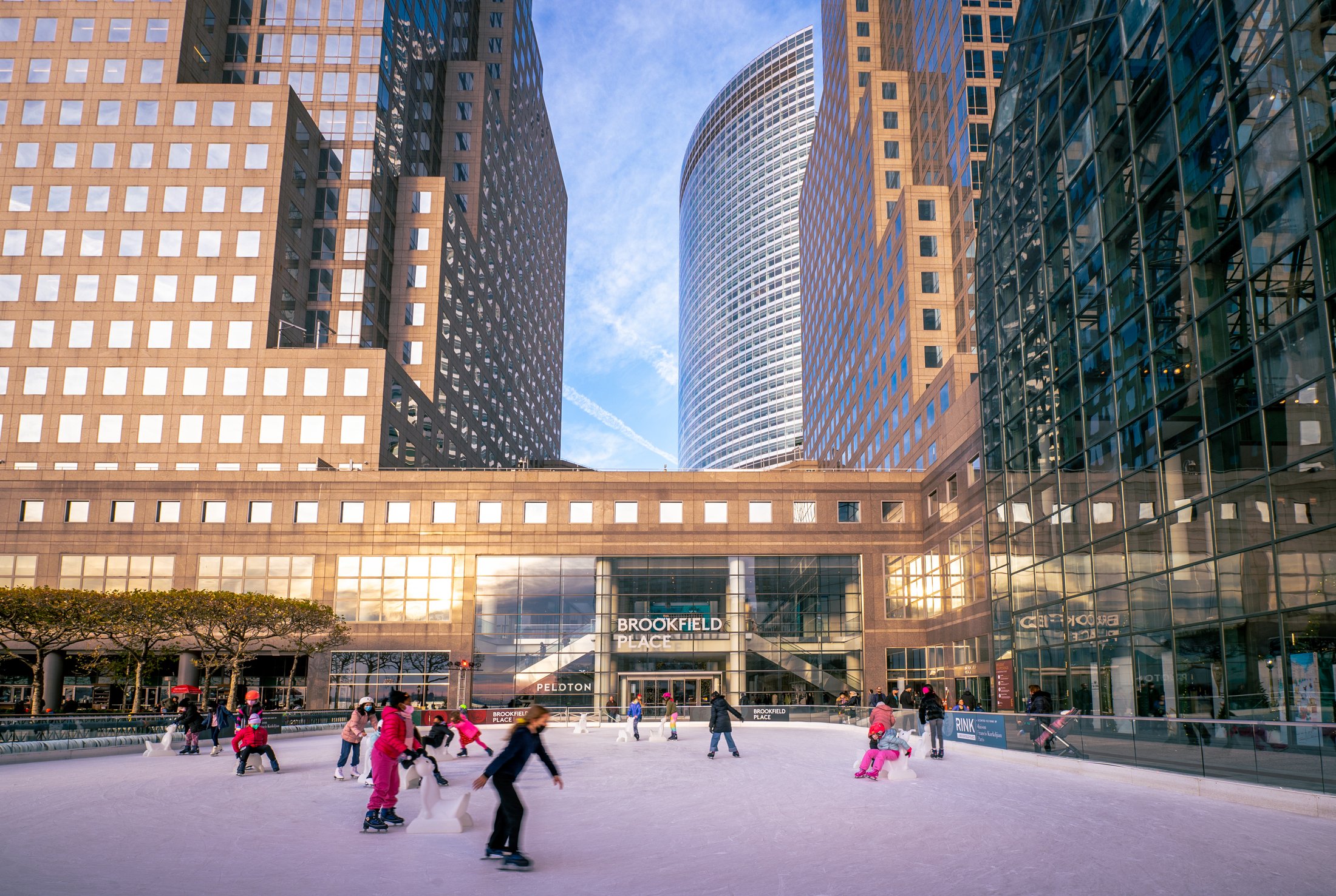 Skaters enjoying an outdoor ice rink near tall, modern glass skyscrapers on a clear day.