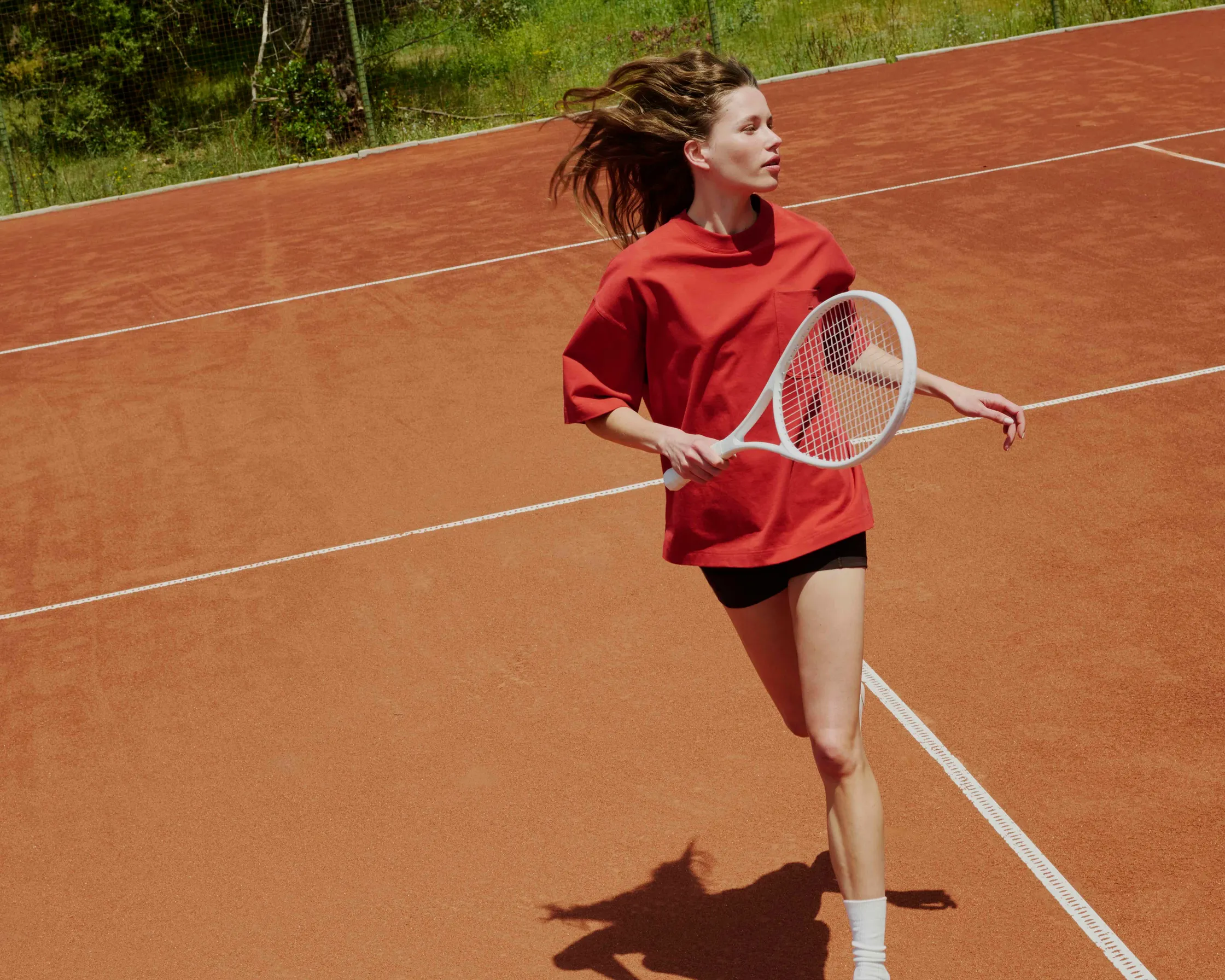 A young woman in a loose red t-shirt and black shorts runs across an outdoor red clay tennis court, holding a white tennis racket.