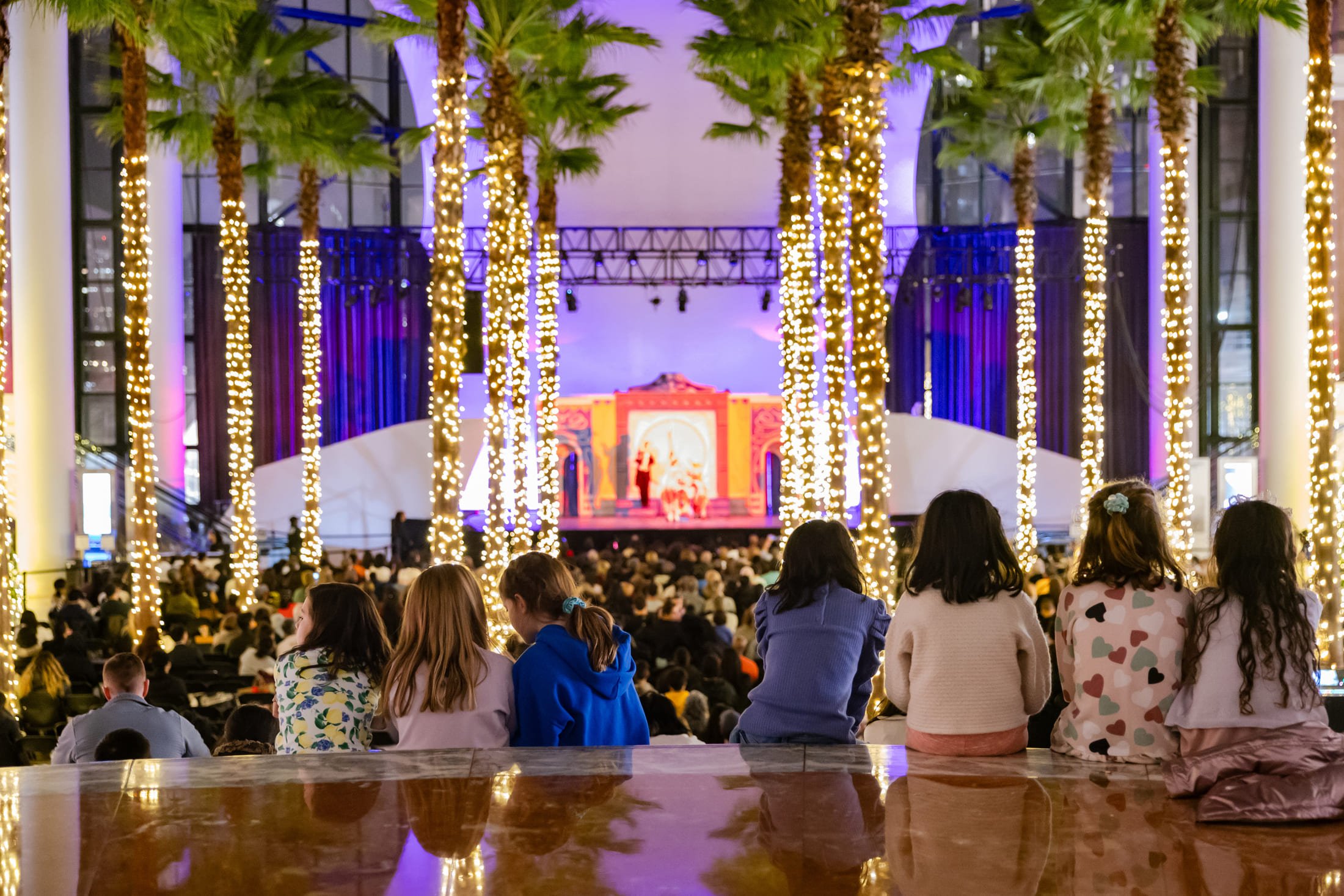 The interior of the Winter Garden at Brookfield Place, showing a crowd watching a performance of the Nutcracker on a stage. Tall palm trees wrapped in strands of white and golden lights frame the audience.