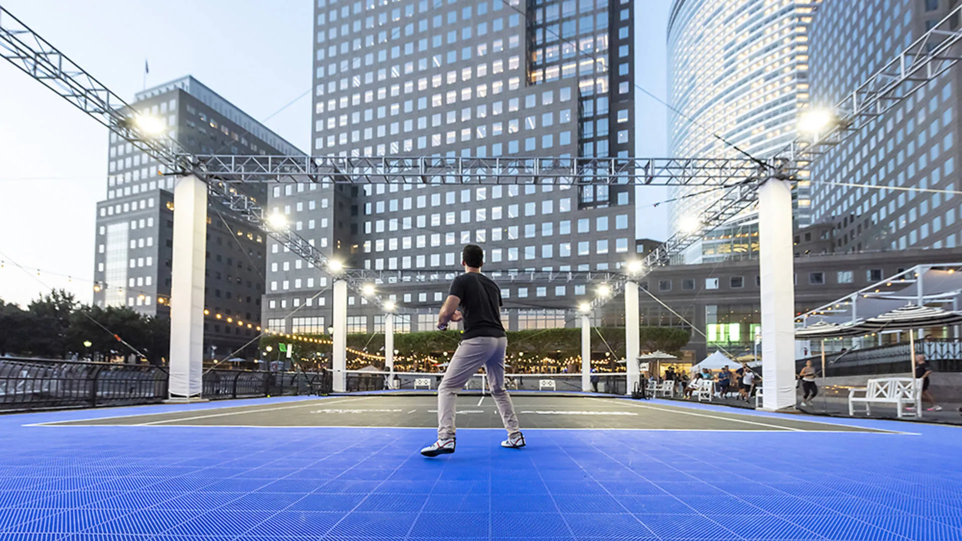 A man stands ready to receive a serve on a blue and gray outdoor tennis court framed by tall modern skyscrapers in the Brookfield Place area.