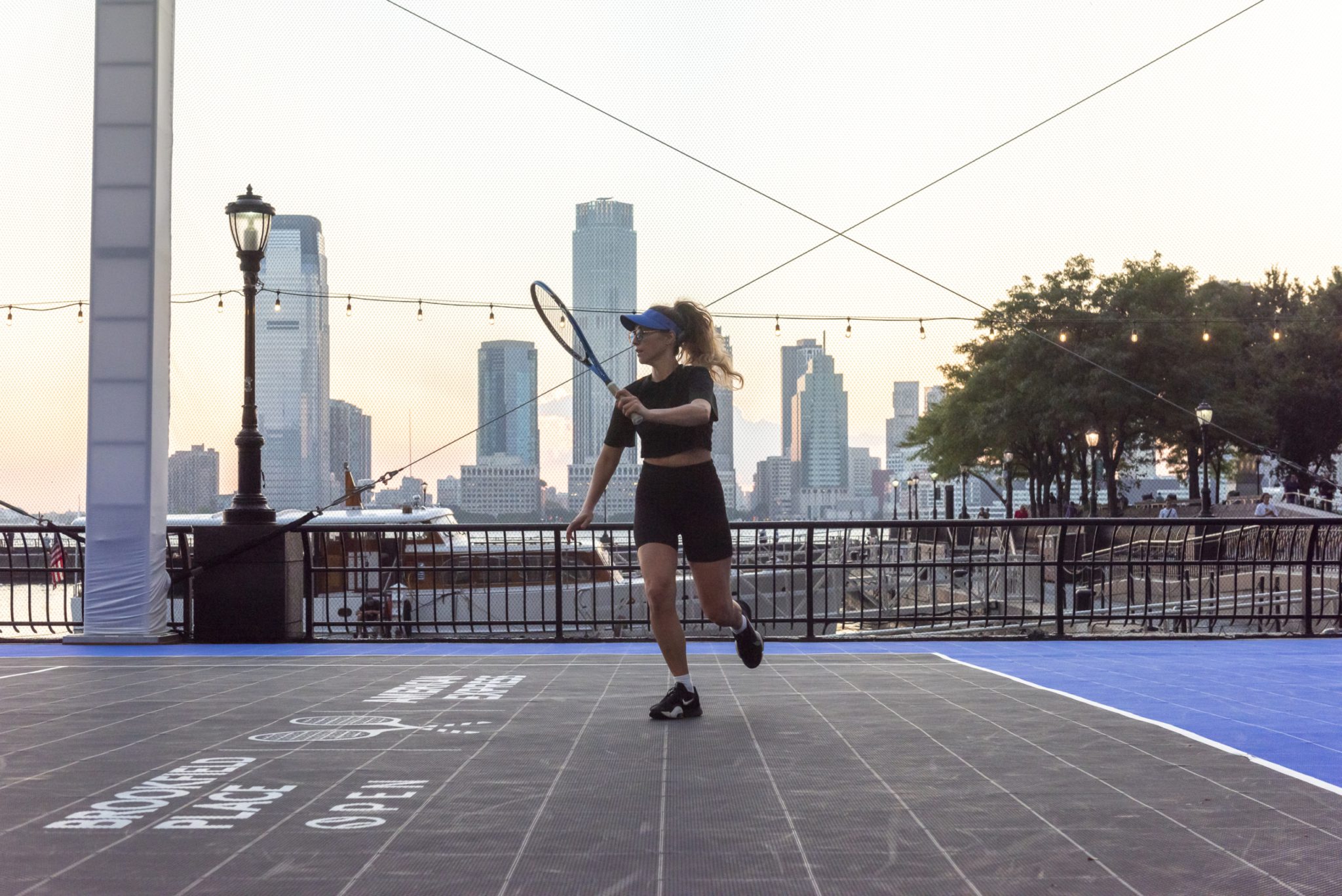 A woman in athletic attire is playing tennis on an outdoor court with the New York City skyline of Jersey City's towers visible in the background at sunset.