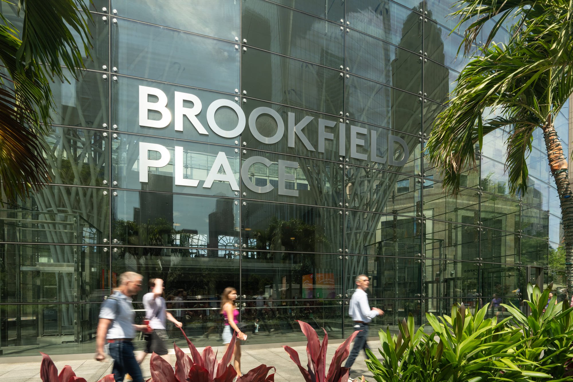 The modern, glass exterior of a Brookfield Place building, featuring the white block text logo on the facade. People are walking along the sidewalk with tropical plants in the foreground.