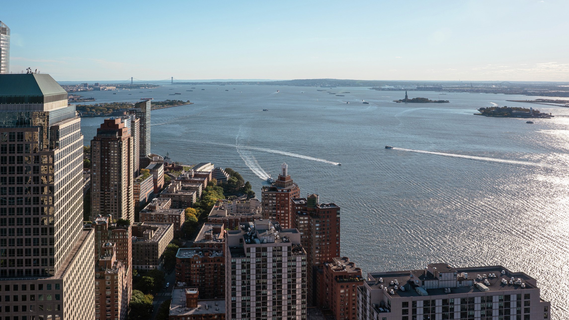 A high-angle, scenic view of the New York Harbor and skyline from a high-rise office building, showing boats on the water, the Statue of Liberty, and the distant bridge.