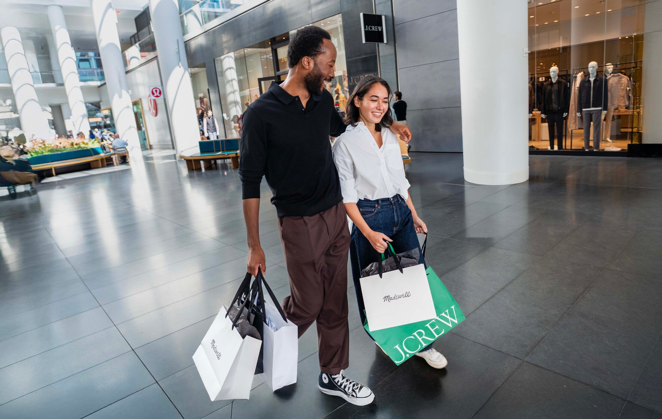 A man and woman walking through the Brookfield Place shopping concourse, smiling and carrying shopping bags from Madewell and J. Crew.