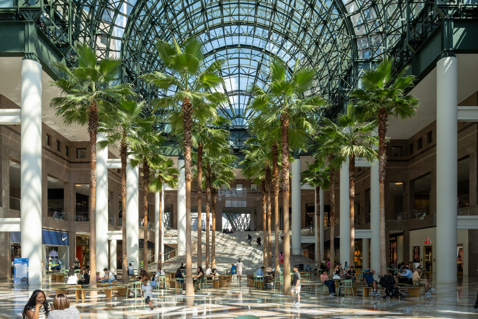 The spacious Winter Garden at Brookfield Place, featuring a soaring, arched glass roof, tall palm trees, and people seated at tables on the marbled floor below.