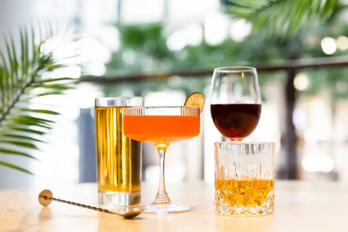 A close-up of four drinks on a marble table in a lounge setting, including a tall yellow drink, an orange cocktail in a coupe glass, red wine, and amber liquor in a rocks glass, framed by palm fronds.